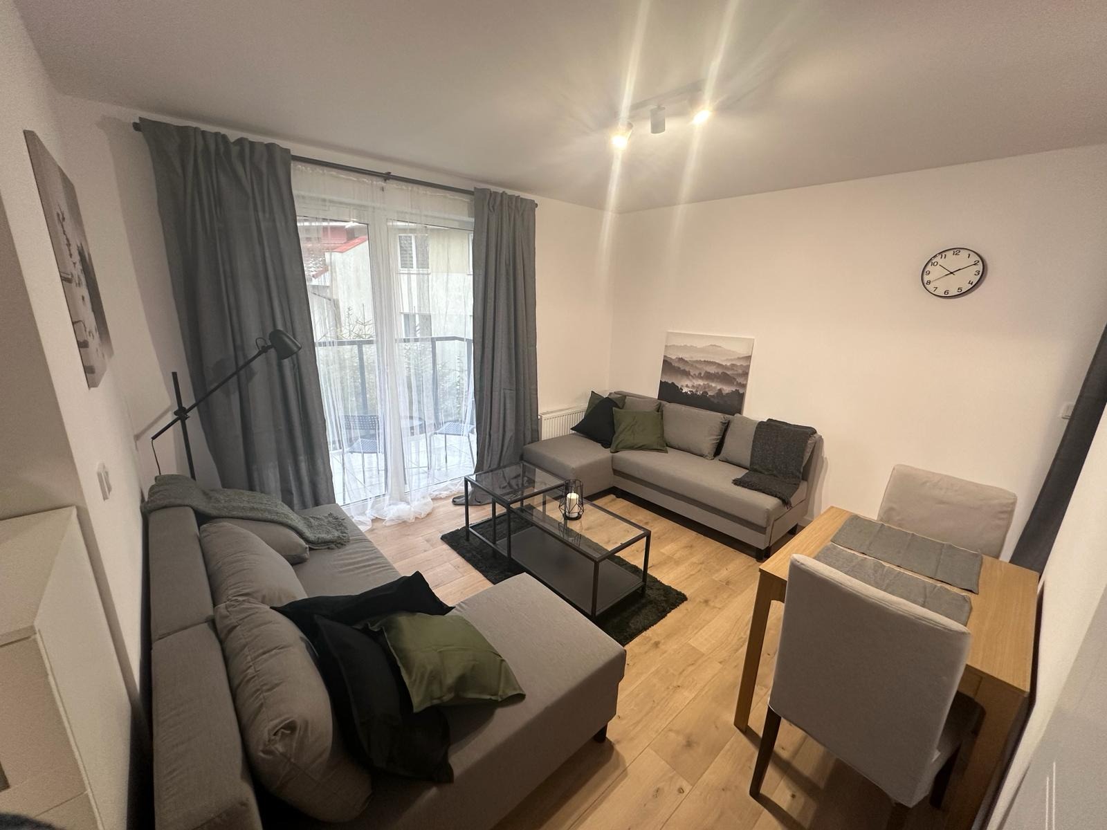 Jasne wnętrze salonu z szarą sofą narożną, stolikiem kawowym i drewnianą podłogą. Widok na balkon przez okno z szarymi zasłonami. Minimalistyczny wystrój.