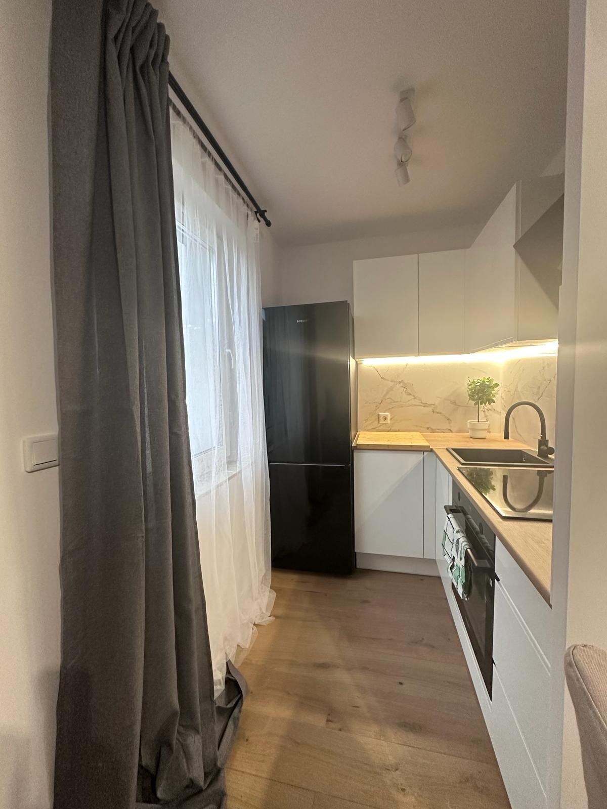 Nowoczesna kuchnia z białymi szafkami, drewnianym blatem i czarną lodówką. Uwagę przyciąga marmurowy backsplash z subtelnym oświetleniem LED.