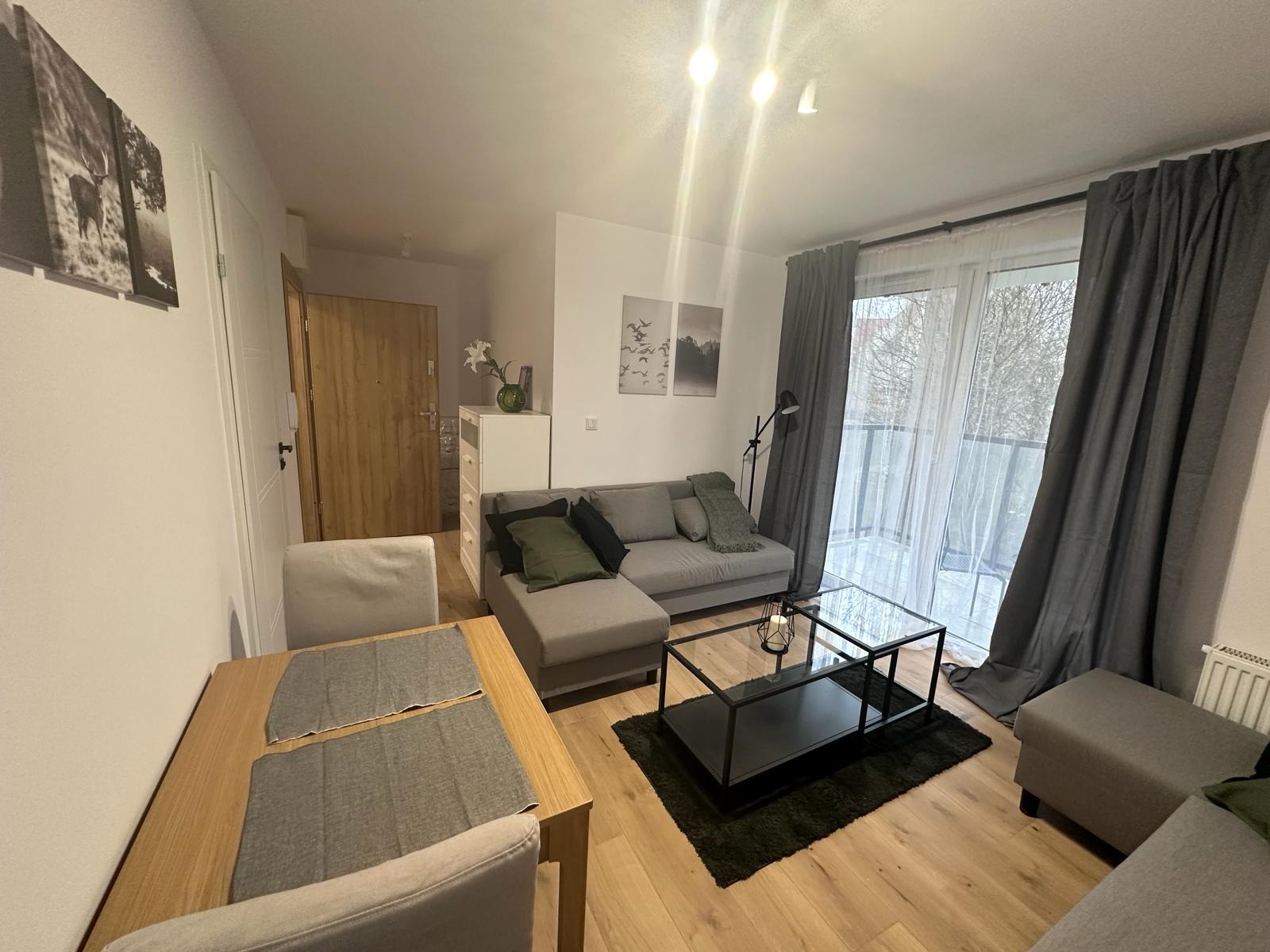 Jasne wnętrze salonu z drewnianą podłogą, szarą sofą narożną, szklanym stolikiem kawowym i zasłonami. Widok na balkon przez okno.