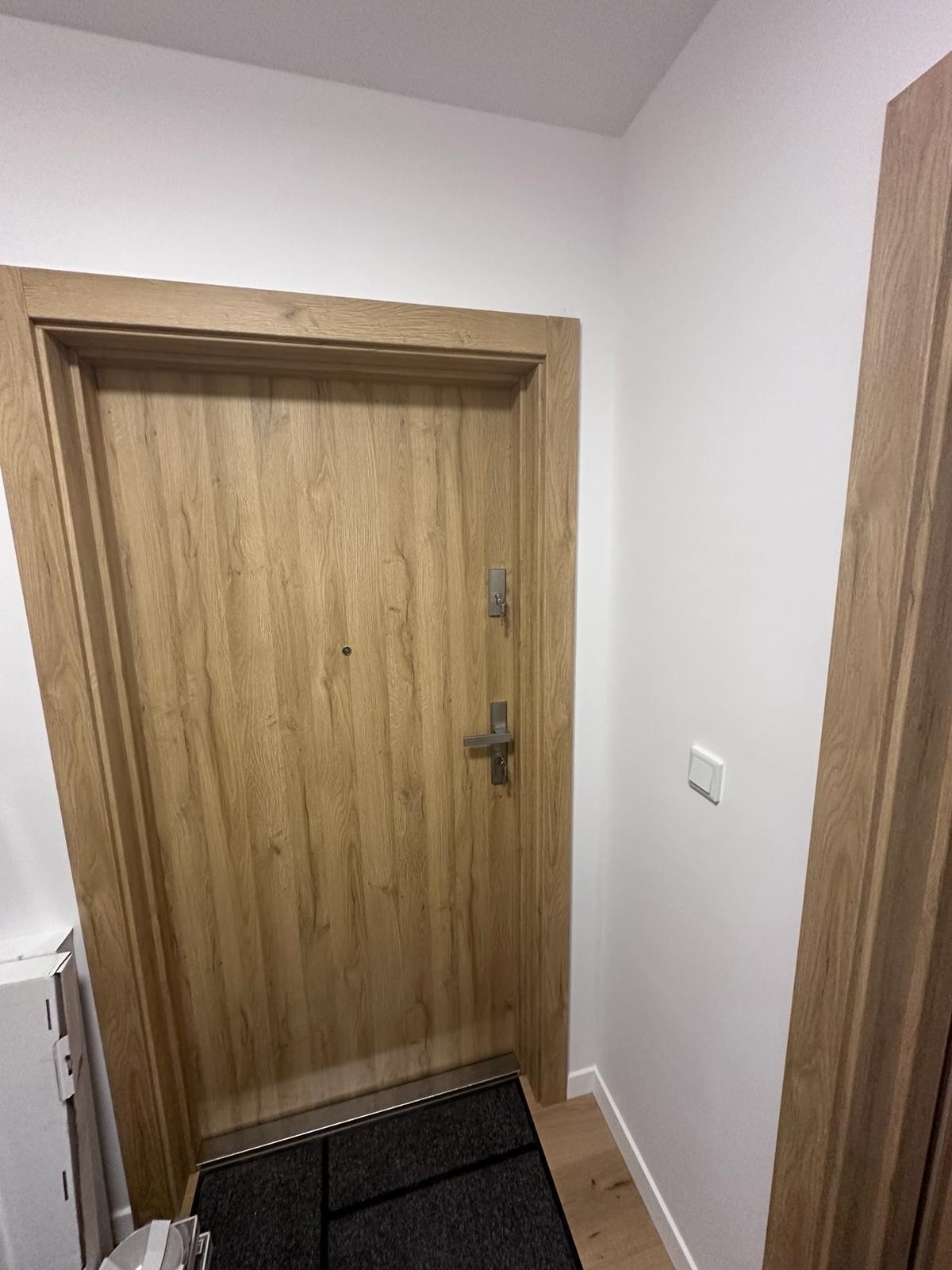 Zamknięte, drewniane drzwi wejściowe z metalową klamką i zamkiem, widoczne od strony wewnętrznej, w jasnym korytarzu z białymi ścianami i drewnianą podłogą.