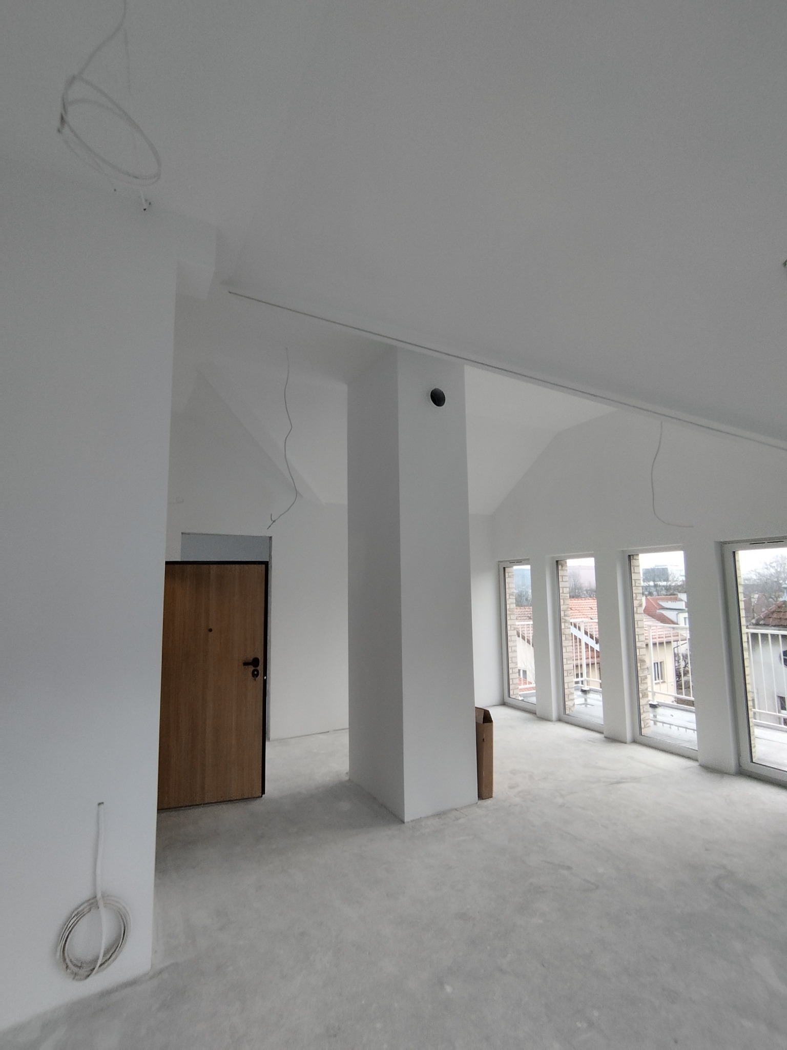 Wnętrze w stanie deweloperskim: białe ściany, surowa podłoga, drewniane drzwi i okna z widokiem na miasto. Widoczne przewody elektryczne i wentylacja.