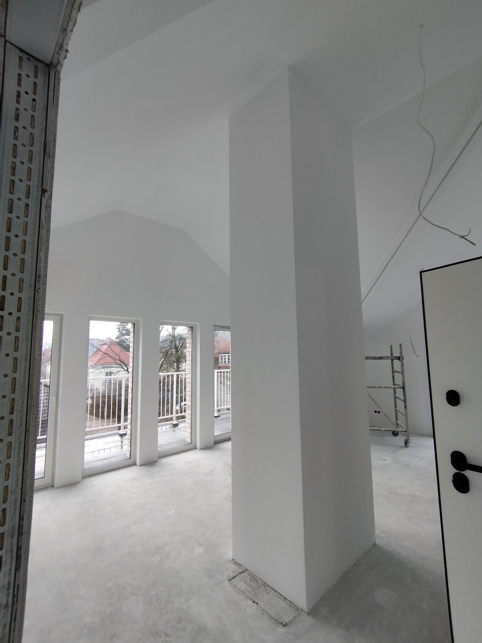 Jasne, przestronne wnętrze w trakcie prac wykończeniowych. Widoczne okna balkonowe, kolumna, surowa podłoga i rusztowanie. Metalowy profil w lewym dolnym rogu.