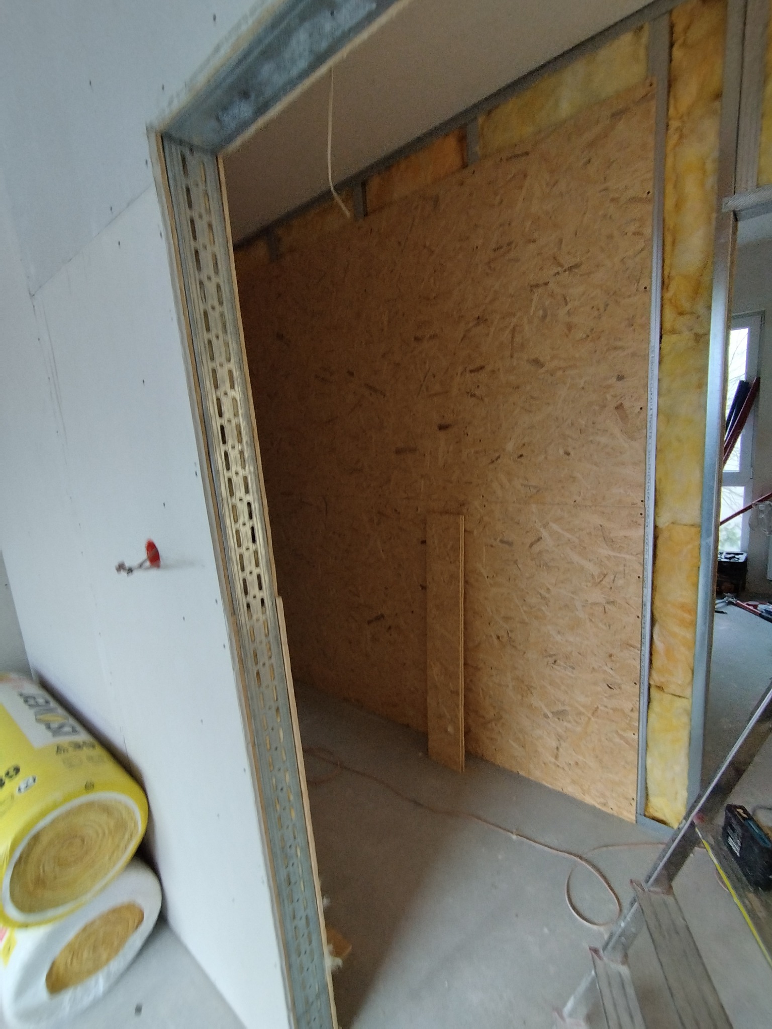 Wnętrze w trakcie prac: płyty OSB na ścianie, izolacja z wełny mineralnej, metalowe profile przy otworze drzwiowym. Widoczne materiały budowlane i drabina.