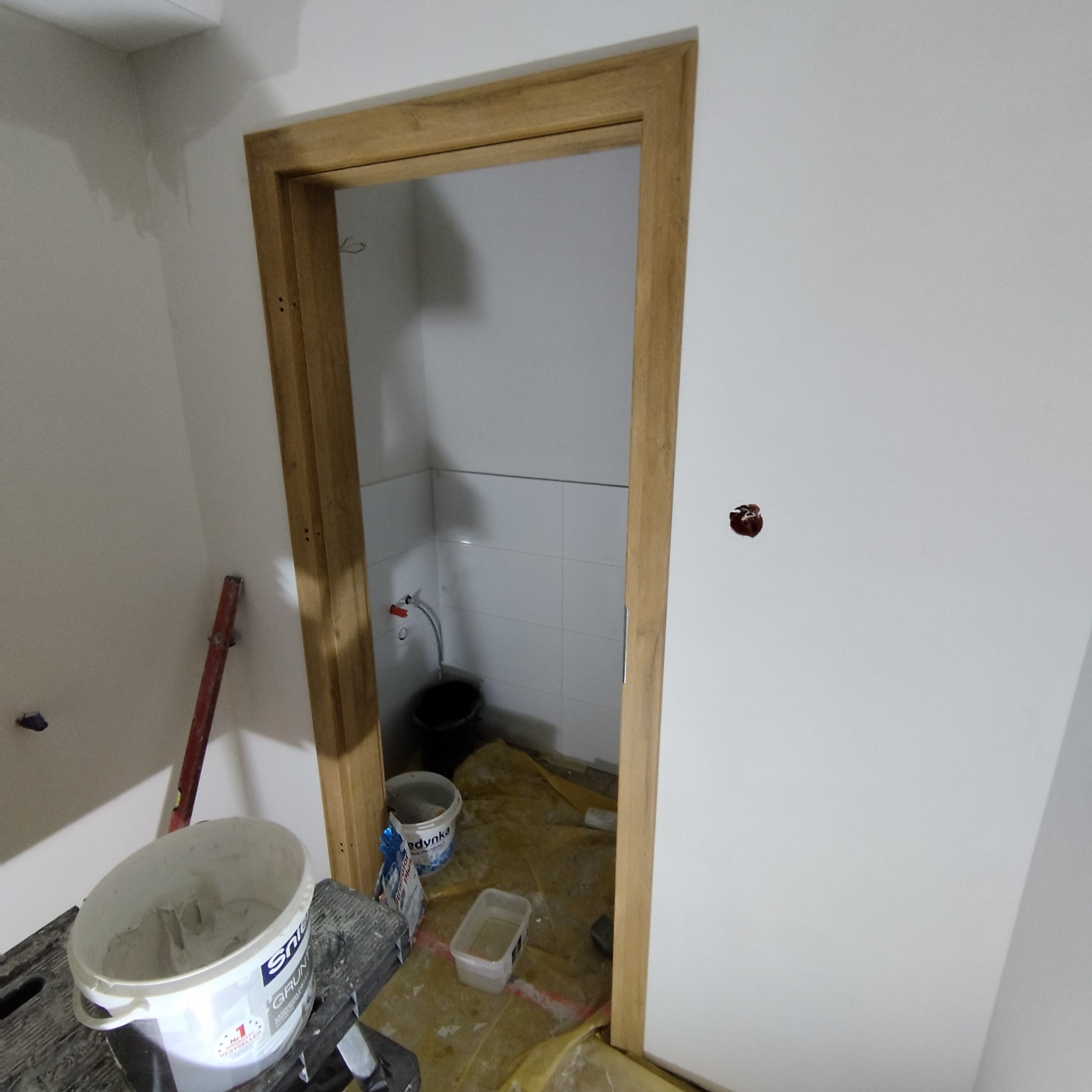 Łazienka w trakcie prac: widoczna drewniana ościeżnica, wiadro z farbą, poziomica i zabezpieczona folią podłoga. Białe płytki na ścianach w tle.