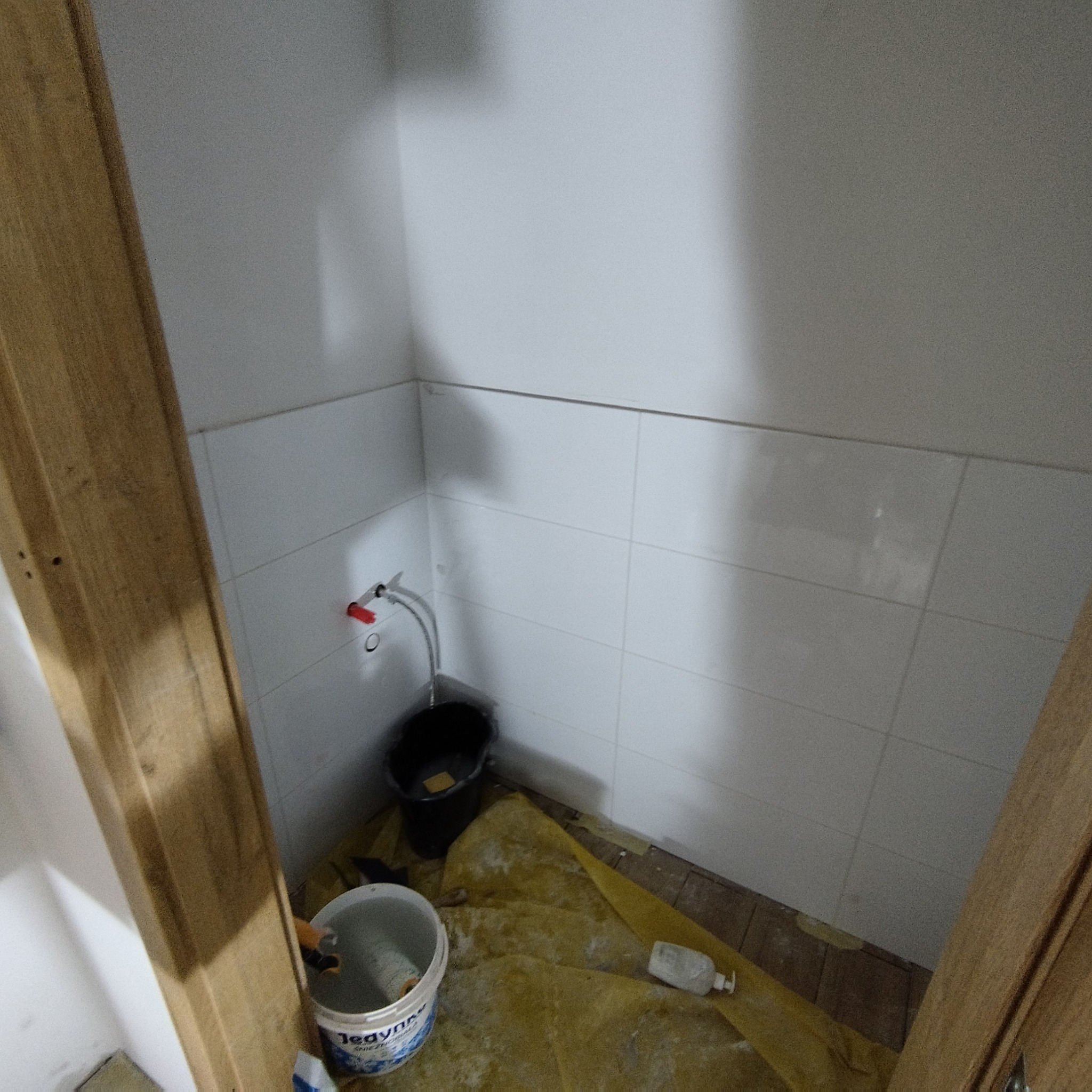 Białe płytki na ścianie w trakcie prac wykończeniowych. Widoczne wiadro z farbą, czarne wiadro i zabezpieczona folią podłoga. Fragment ościeżnicy po lewej i prawej stronie.