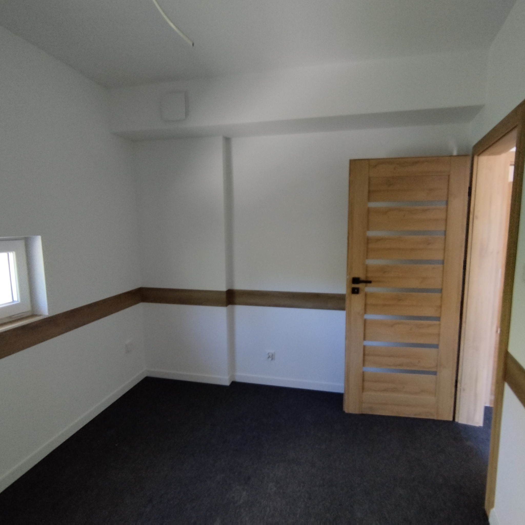 Jasne wnętrze po odświeżeniu: białe ściany z drewnianą listwą, ciemna podłoga i drewniane drzwi z przeszkleniami. Widok na otwarte przejście do kolejnego pomieszczenia.