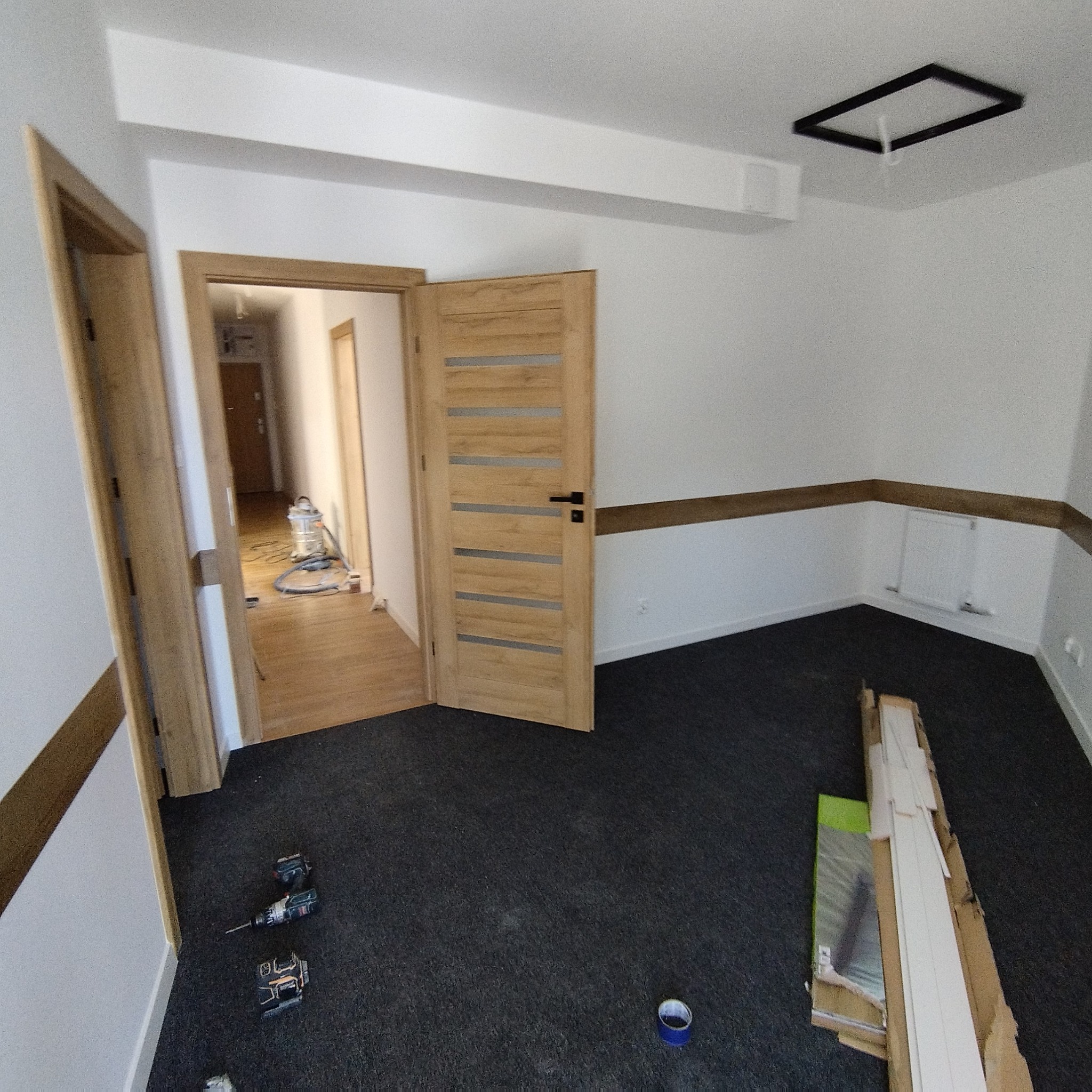 Wnętrze pokoju z dwiema otwartymi drewnianymi drzwiami, narzędziami na podłodze i listwami przypodłogowymi w opakowaniu. Widoczny fragment sufitu z lampą.