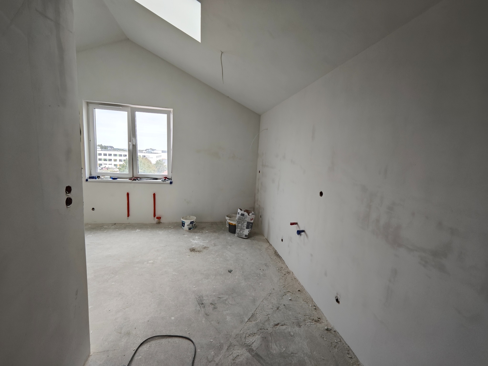Surowe wnętrze poddasza z oknem, widoczne ślady szpachlowania na ścianach, przygotowane instalacje wodne i elektryczne, worki z zaprawą budowlaną.