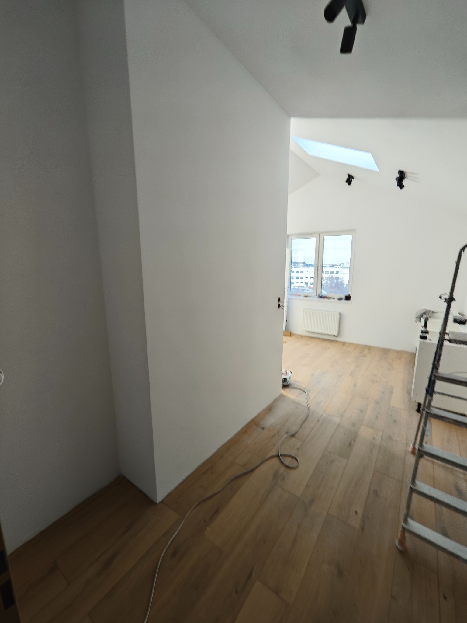 Jasne wnętrze po odświeżeniu: drewniana podłoga, białe ściany, okno dachowe. Widoczna drabina i narzędzia, sugerujące trwające prace wykończeniowe.