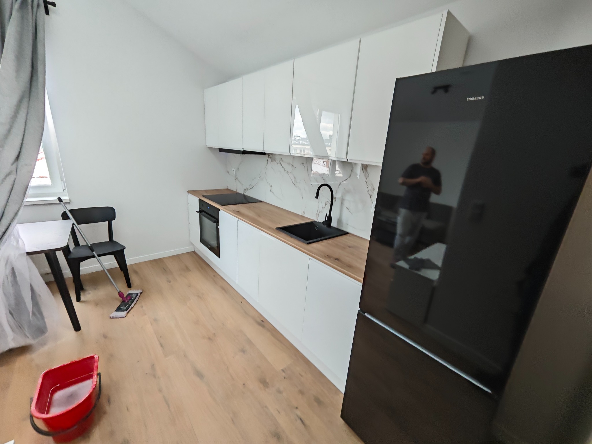 Nowoczesna, jasna kuchnia z białymi szafkami i drewnianym blatem. Widoczny czarny zlew, płyta grzewcza i lodówka. Na podłodze mop i wiadro, co sugeruje sprzątanie po remoncie.