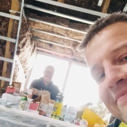 Daniel Wawrzyniak - Montaż sufitu z płyt karton-gips: widok z dołu na konstrukcję metalową, izolację z wełny mineralnej i dwóch monterów podczas pracy. Ujęcie z bliska, z perspektywy jednego z nich.