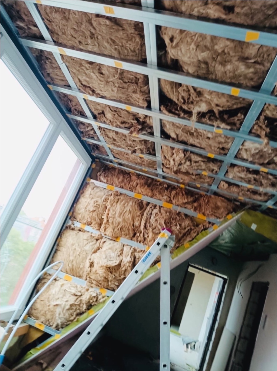 Skośny sufit poddasza z izolacją z wełny mineralnej, widoczne metalowe profile i drabina Krause. Prace wykończeniowe w trakcie realizacji.