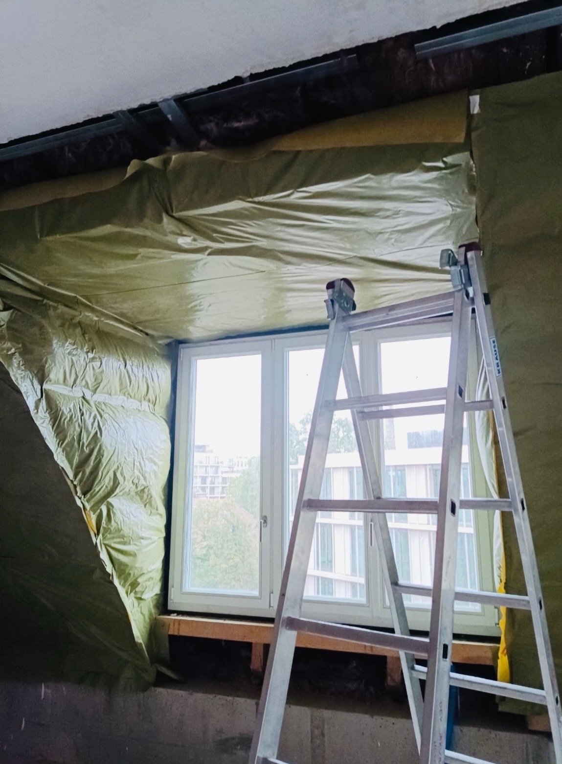 Izolacja poddasza wełną mineralną, zabezpieczoną folią, widoczna drabina aluminiowa i okno. Prace wykończeniowe w trakcie realizacji.