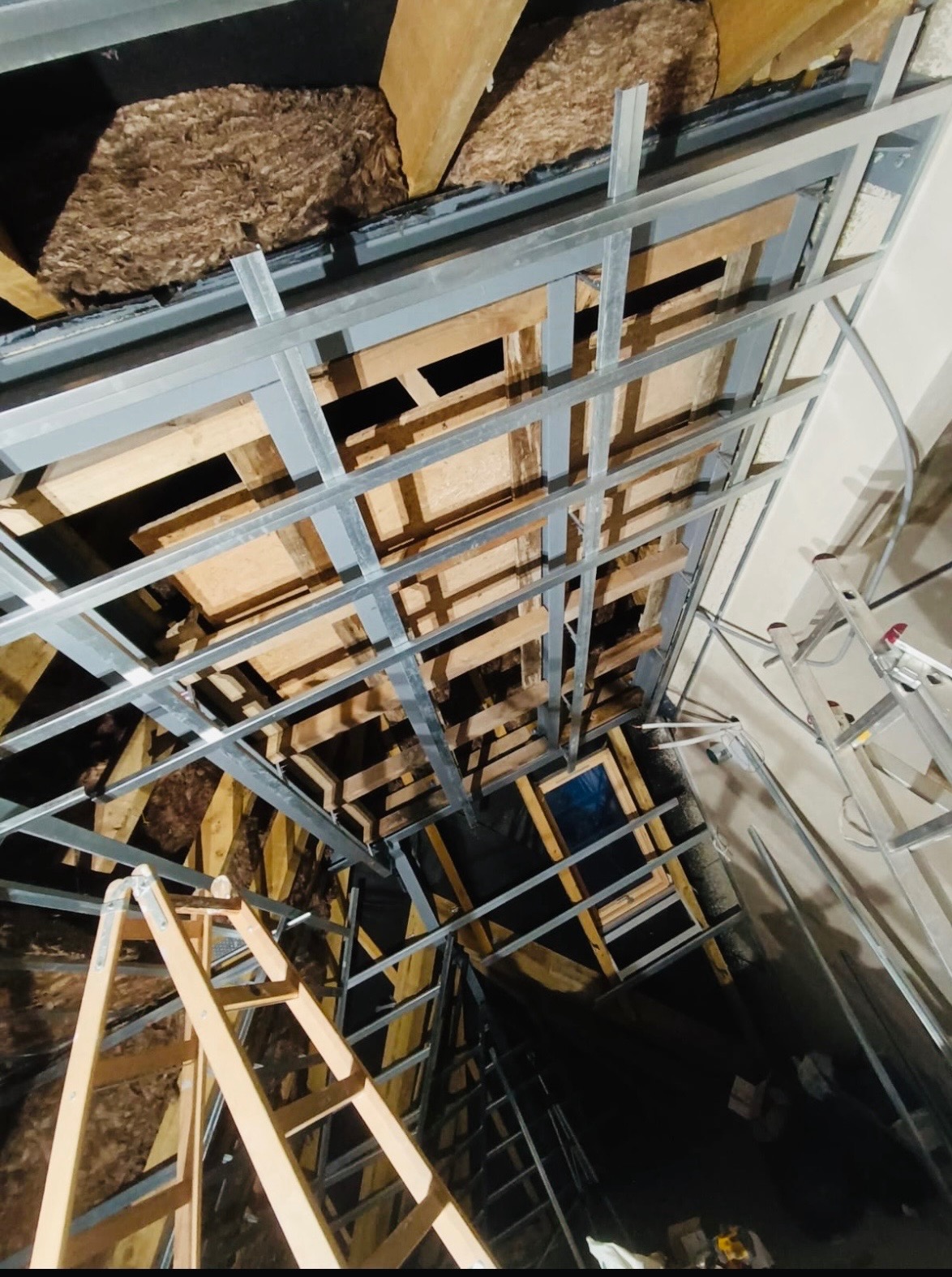 Sufit podwieszany w trakcie montażu: metalowa konstrukcja, izolacja z wełny mineralnej, drewniane elementy. Widok z dołu, drabina w kadrze.