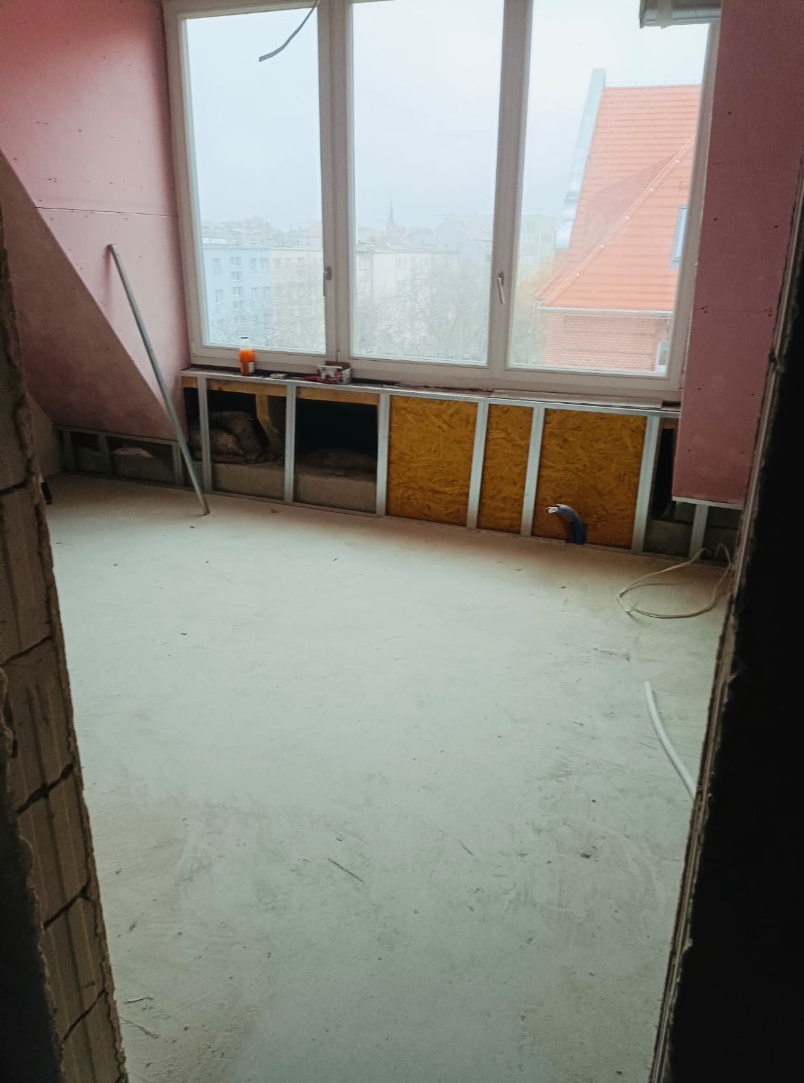 Poddasze w trakcie adaptacji: ściany z płyt karton-gips, okno z widokiem na miasto, zabudowa pod oknem z widocznymi elementami instalacji. Prace wykończeniowe.