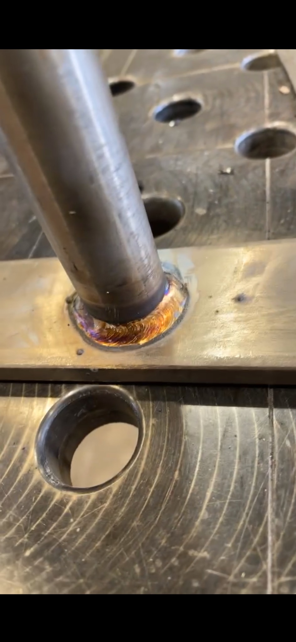 Metalowa rura spawana do płyty z otworami, widoczne tęczowe przebarwienia na spoinie, co świadczy o wysokiej temperaturze i jakości wykonania.