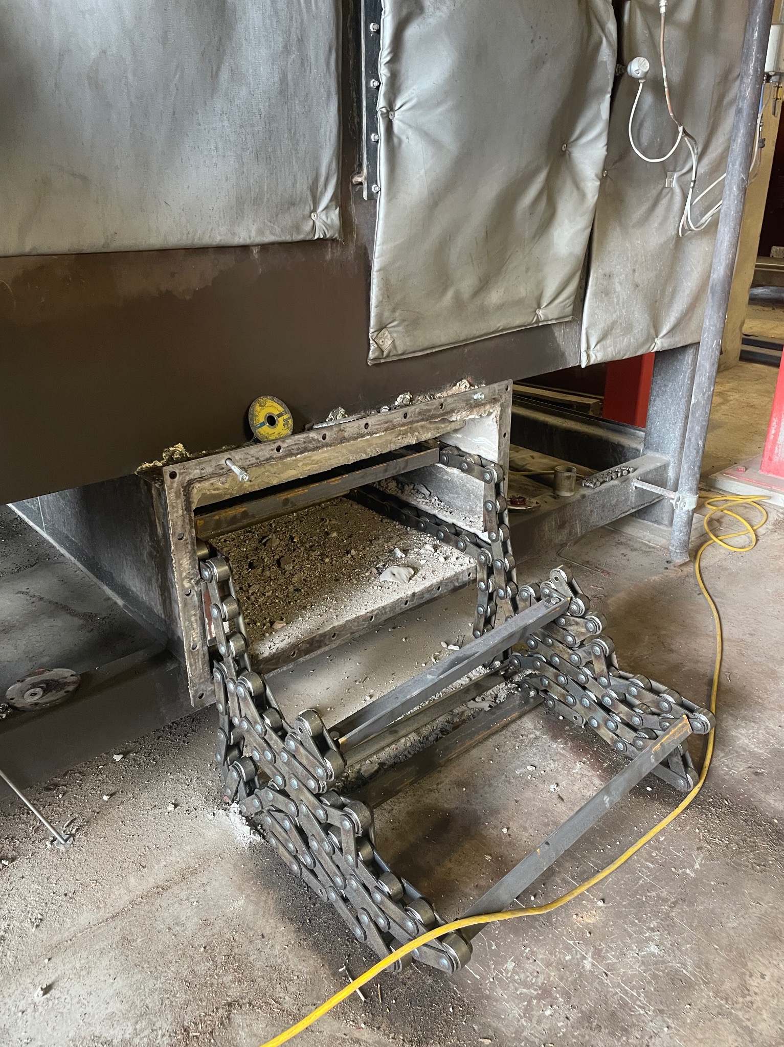 Metalowa konstrukcja schodkowa z łańcucha, przymocowana do otworu w brązowej obudowie. Widoczne ślady spawania i żółty kabel na podłodze.