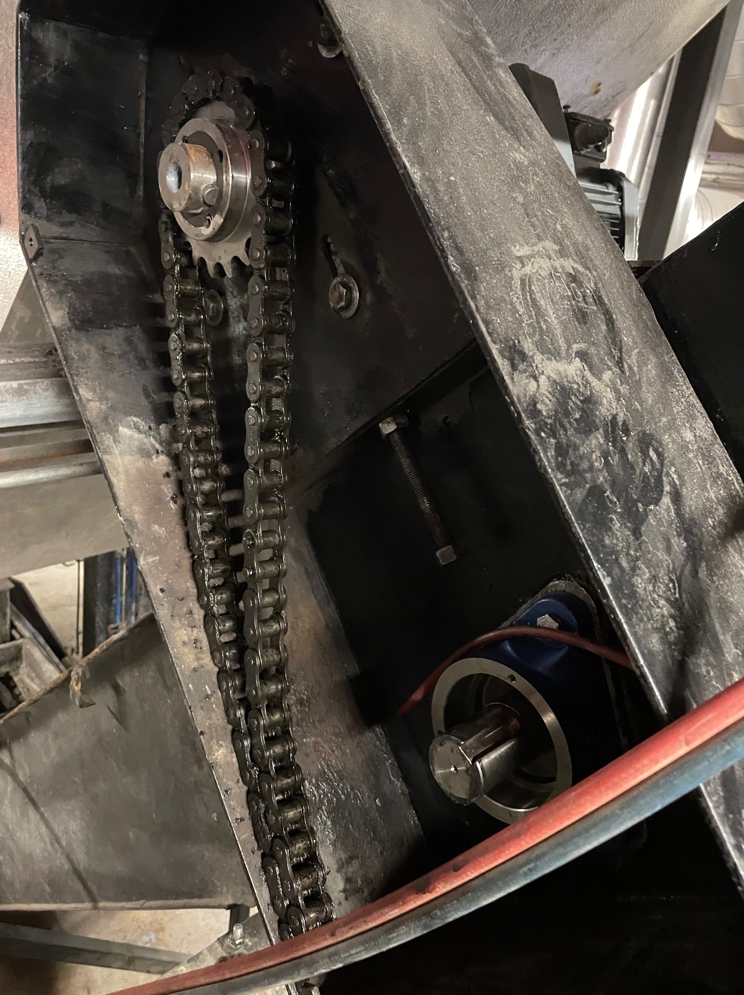 Metalowa konstrukcja z łańcuchem i zębatką, widoczne elementy silnika. Detal mechanizmu w hali produkcyjnej, oświetlenie górne.