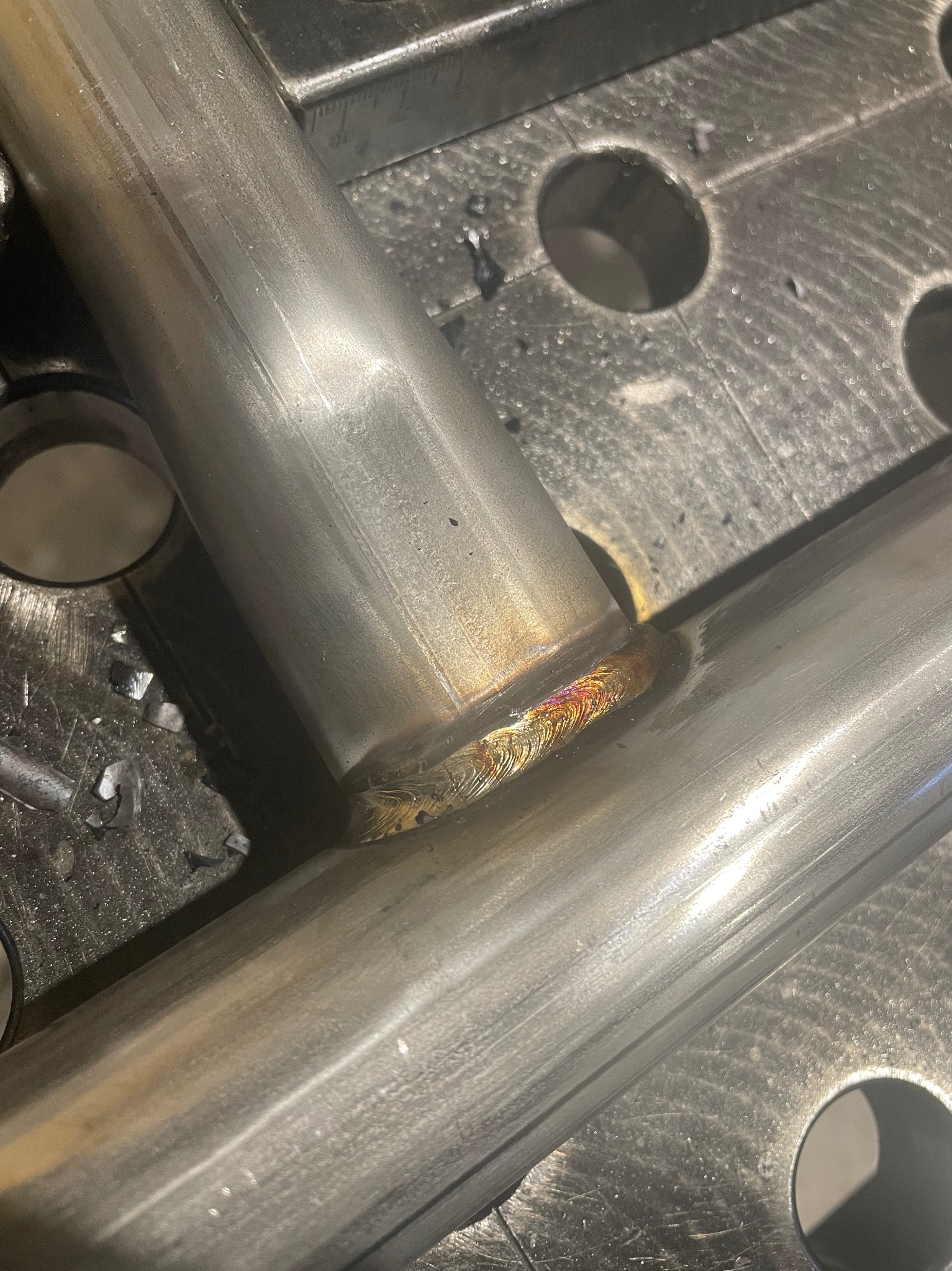 Spoina łącząca dwie metalowe rury na perforowanym stole spawalniczym. Widoczne ślady obróbki i opiłki metalu. Charakterystyczne zabarwienie spoiny.