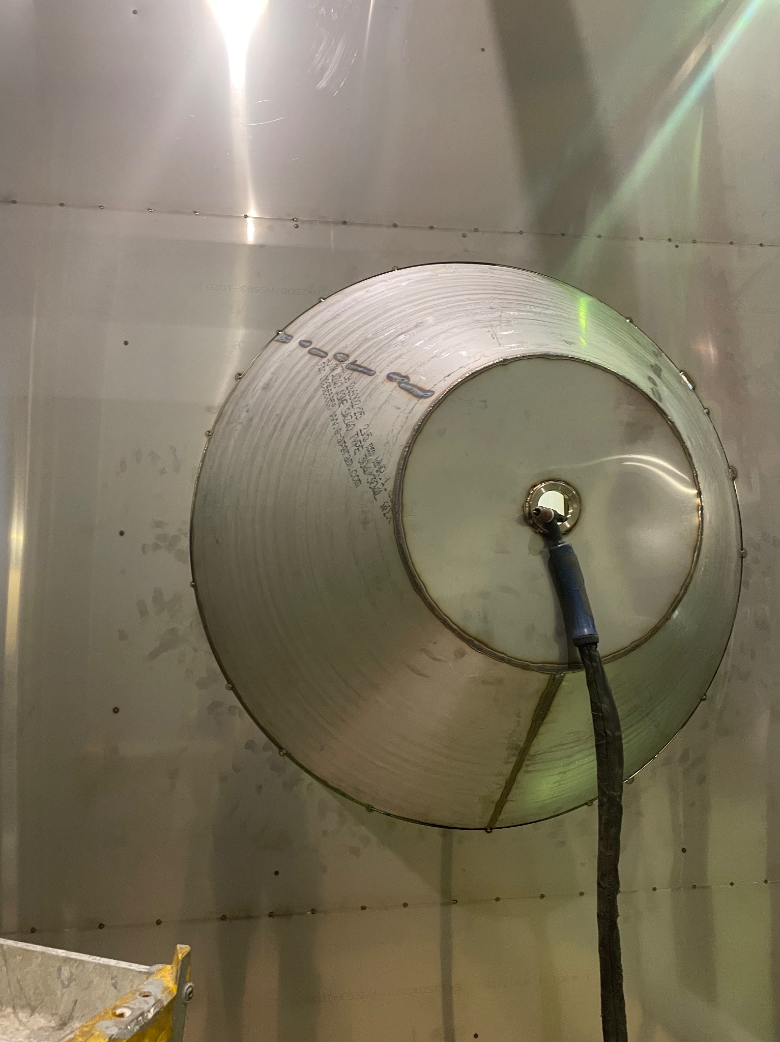 Spawanie zbiornika ze stali nierdzewnej metodą TIG, widoczne ślady spawania i oznaczenia na metalu. Kabel spawalniczy w centrum kadru, oświetlenie warsztatowe.