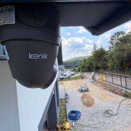 LOOK CONNECT Kacper Kwaśniowski - Ciemnoszara kamera bezpieczeństwa marki Kenik, zamontowana pod okapem budynku, z widokiem na plac budowy z układaną kostką brukową, narzędziami i pracownikiem w tle.