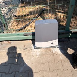 LOOK CONNECT Kacper Kwaśniowski - Szary napęd do bramy przesuwnej CAME IT, zamontowany na betonowej podstawie obok zielonego ogrodzenia panelowego. Widoczne zębate listwy i cień osoby z telefonem na kostce brukowej.