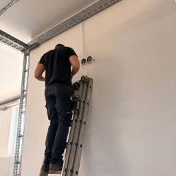 LOOK CONNECT Kacper Kwaśniowski - Technik w Budzowie montuje elementy instalacji na białej ścianie, stojąc na drabinie. Widoczne metalowe korytka kablowe na suficie i pionowo, narzędzia przy pasie.