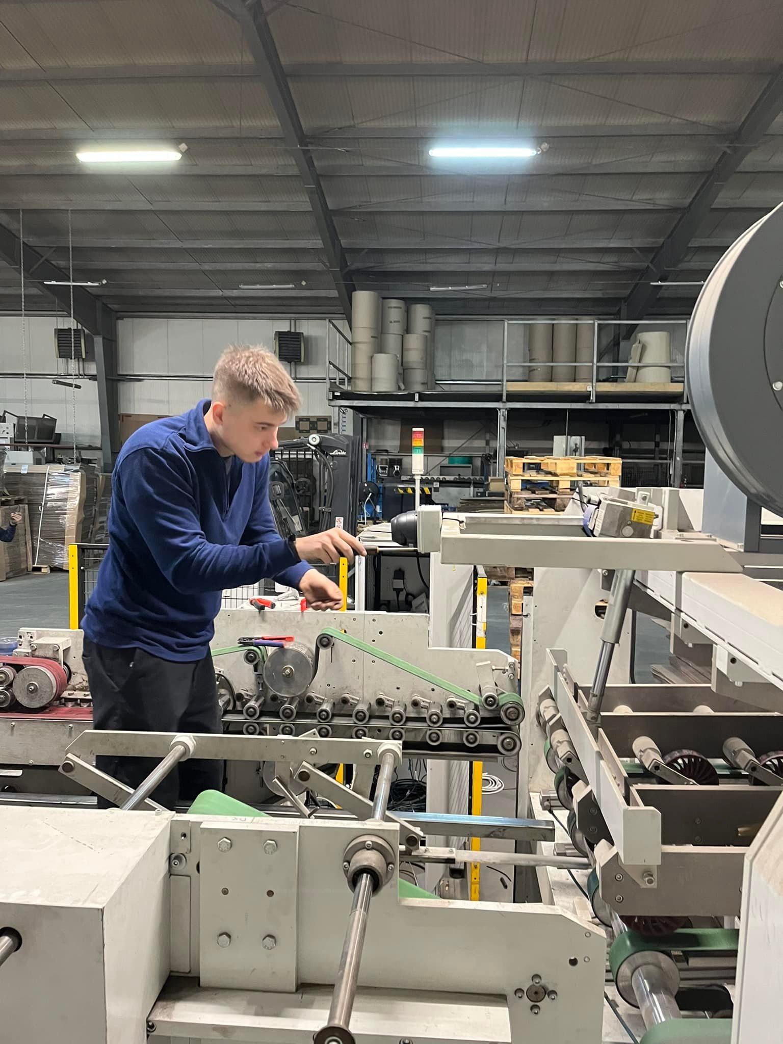Pracownik w niebieskiej bluzie obsługuje maszynę przemysłową w hali produkcyjnej. Widoczne rolki, pasy transmisyjne i elementy sterujące. Oświetlenie jarzeniowe.