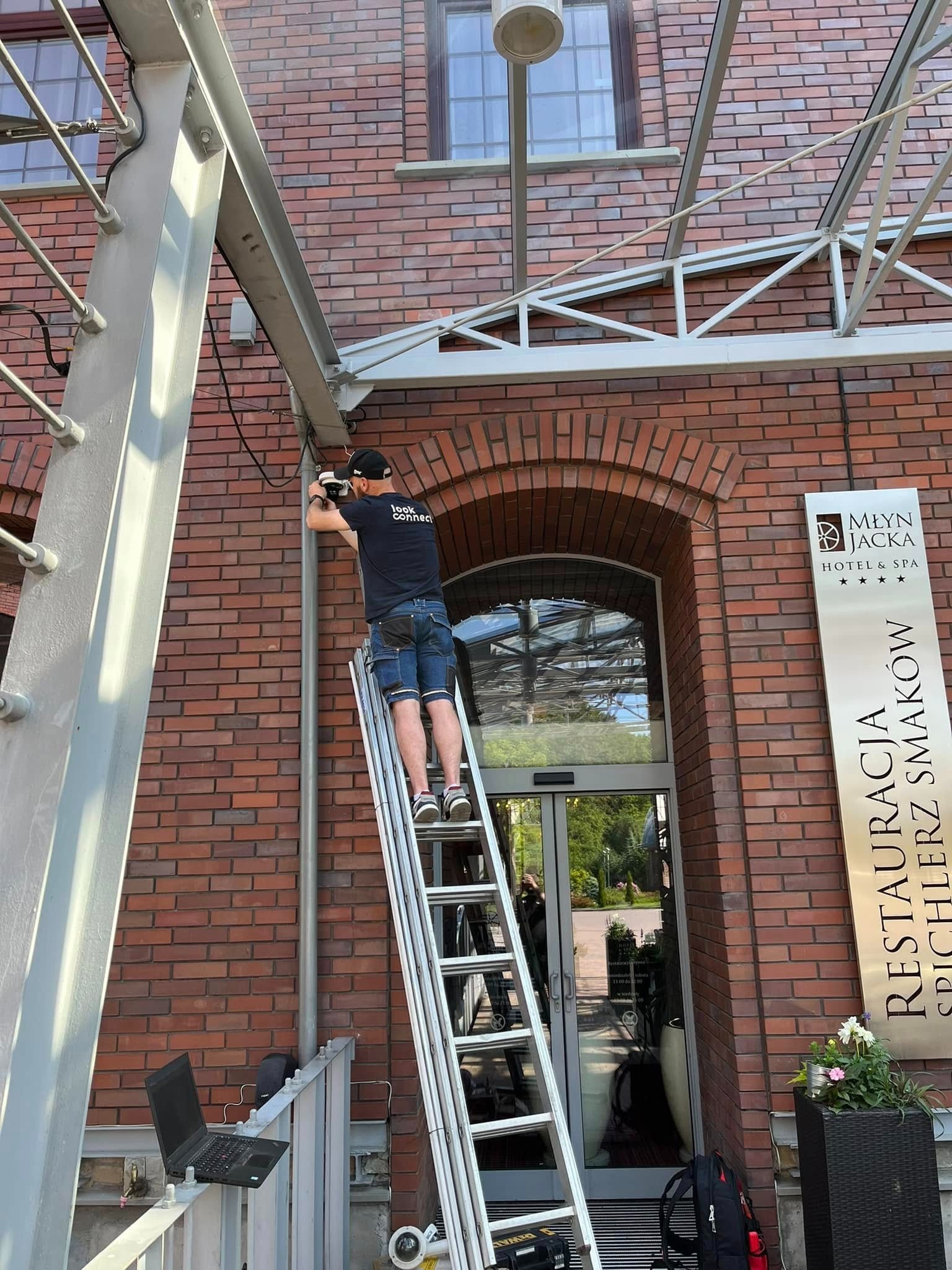 Instalator na drabinie montuje kamerę monitoringu na ceglanej fasadzie budynku Młyn Jacka Hotel & SPA. Widoczny laptop i narzędzia.