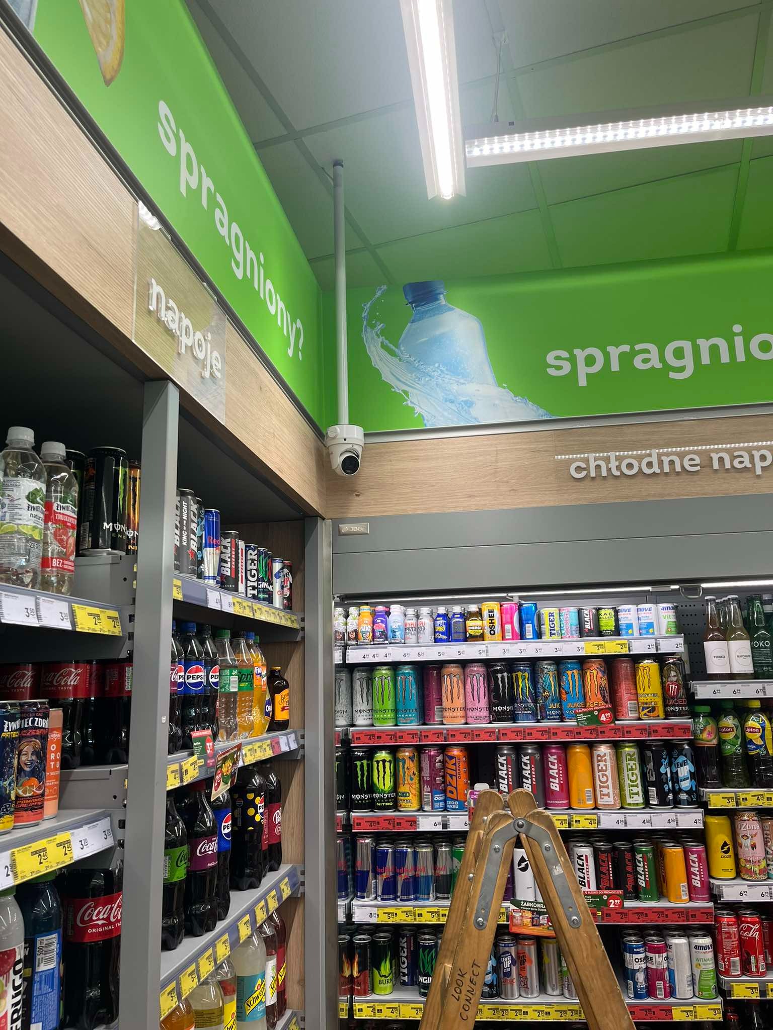 Kamera monitoringu dyskretnie zamontowana w sklepie spożywczym, nad regałami z napojami, zapewniająca bezpieczeństwo i nadzór nad asortymentem.