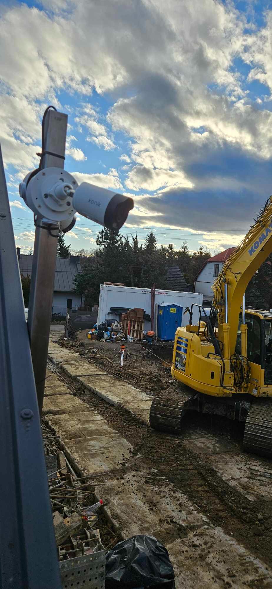 Kamera Kenik na słupie, z tyłu plac budowy z koparką Komatsu, kontenerem i przenośną toaletą. Błękitne niebo z chmurami w tle. Ujęcie z dołu, z lewej strony kadru.