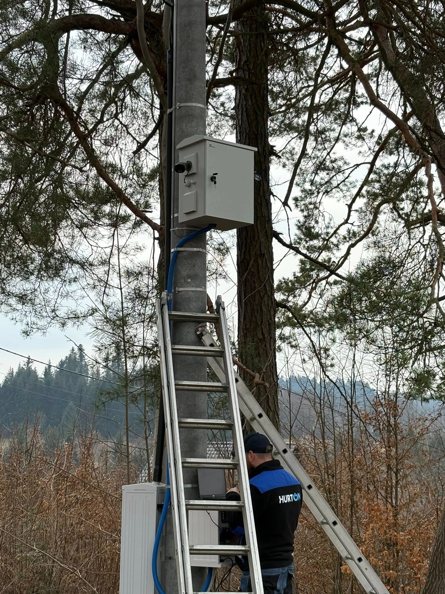 Instalacja kamery monitoringu na słupie przez montera na drabinie. Widoczna puszka z elektroniką i niebieski peszel. W tle drzewa i las.