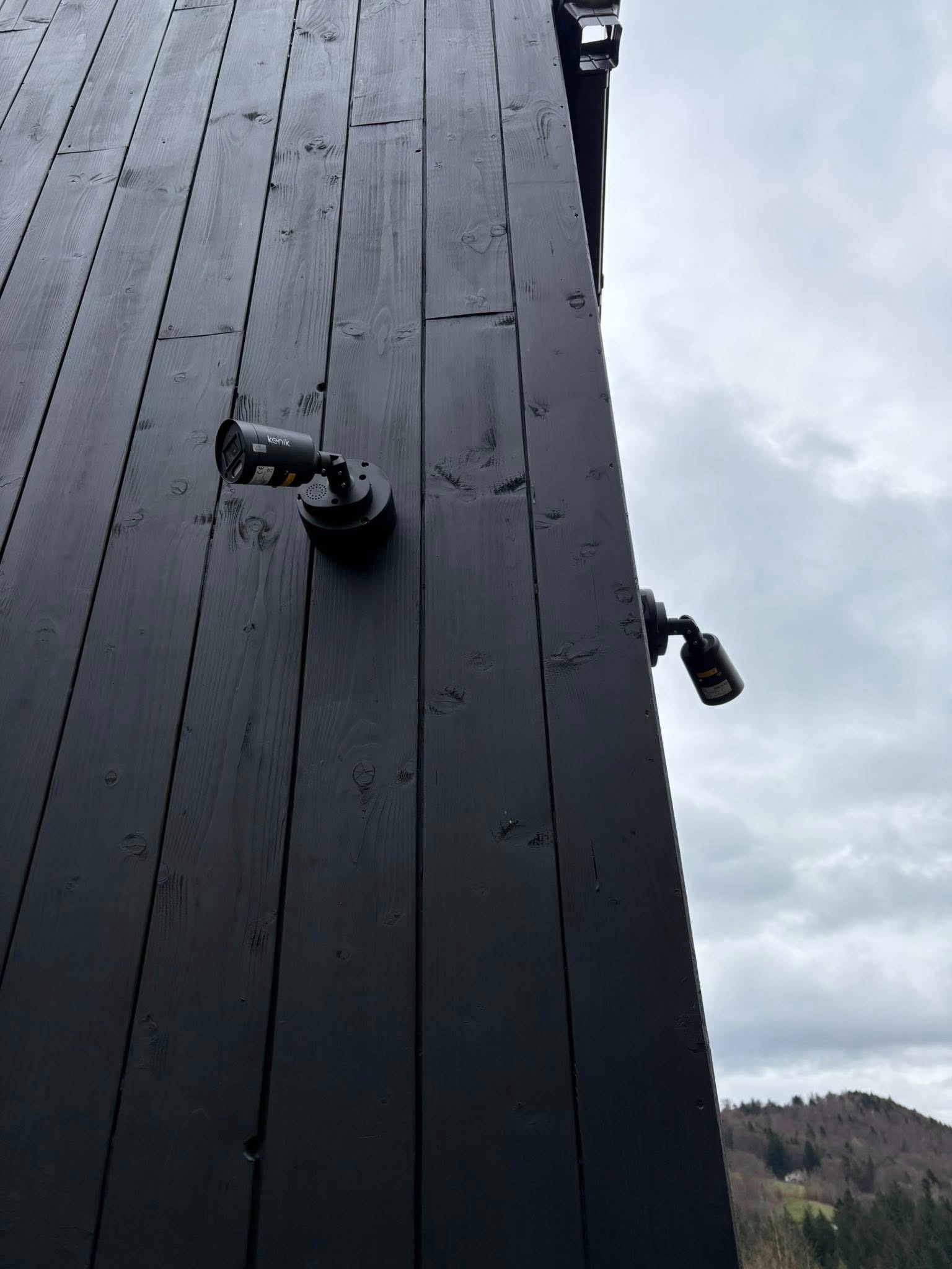 Dwie kamery monitoringu Kenik zamontowane na czarnej, drewnianej elewacji budynku. Widok pod kątem, w tle pochmurne niebo i zalesione wzgórze.