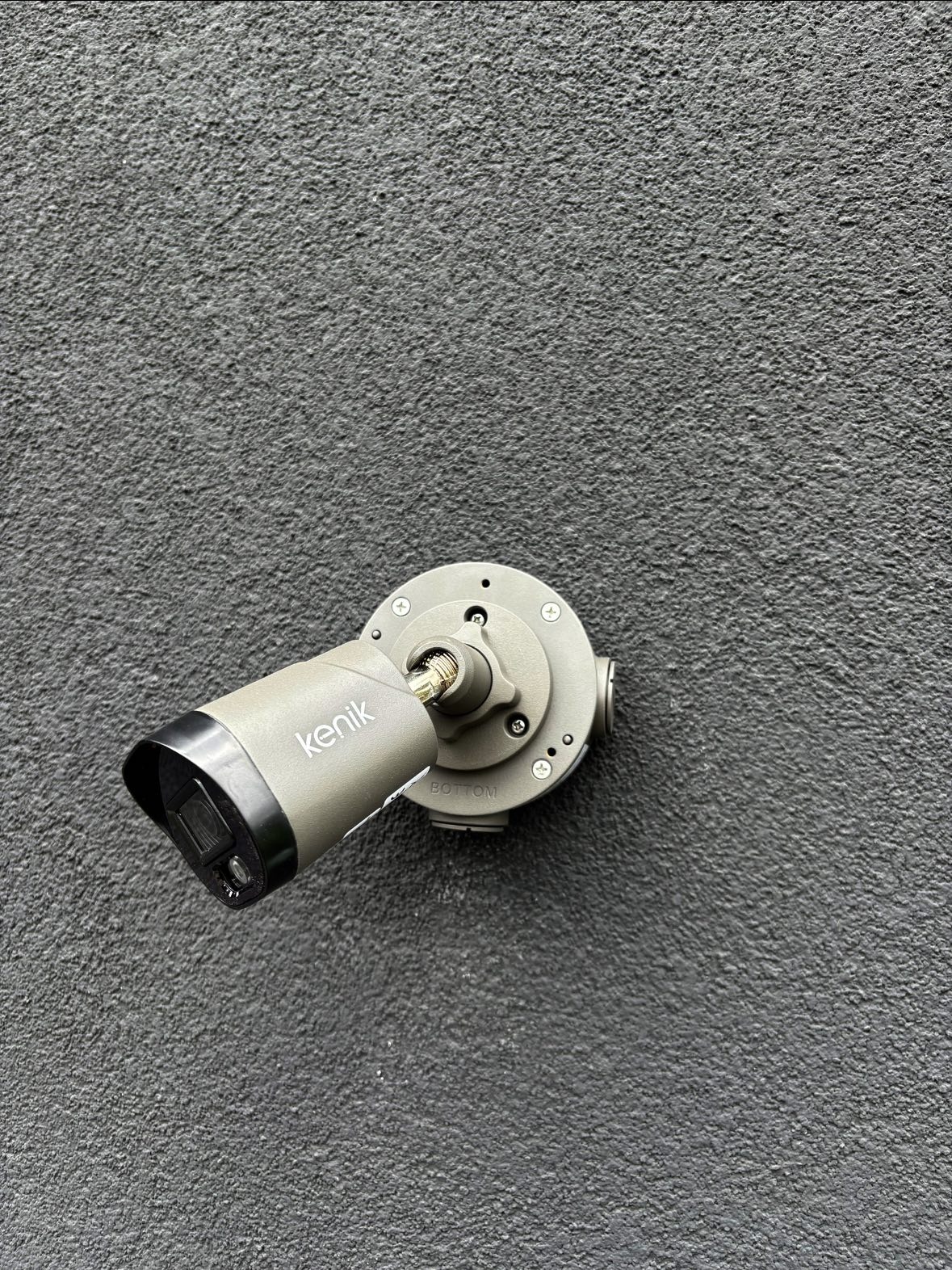 Szara kamera monitoringu marki Kenik zamontowana na szarej, teksturowanej ścianie. Widoczny uchwyt i obiektyw kamery. Kąt ujęcia z dołu.