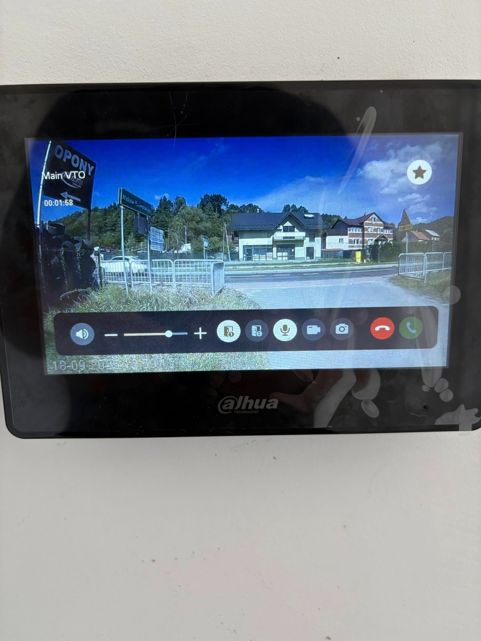 Ekran wideodomofonu Dahua prezentujący obraz z kamery na zewnątrz, z widocznymi budynkami i elementami infrastruktury. Interfejs użytkownika z ikonami połączeń i regulacji głośności.