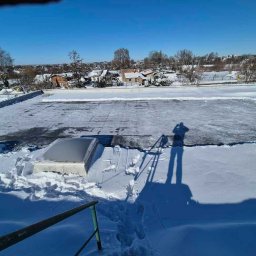 Śnieg na dachu budynku z świetlikiem, cień osoby na śniegu, widok na zasypane śniegiem domy i drzewa w oddali, błękitne niebo.