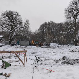 Krzysztof Krankowski - Zimowy krajobraz z wykopem, traktorem i stertami śniegu. Widoczne drzewa bez liści i budynek w tle. Prace ziemne w trakcie odśnieżania.