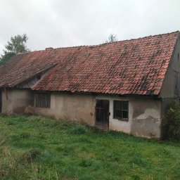 Krzysztof Krankowski - Stary, zaniedbany budynek z czerwoną dachówką, porośnięty trawą. Widoczne okna i drzwi, ściany z ubytkami tynku. Częściowo widoczna osoba po lewej stronie kadru.