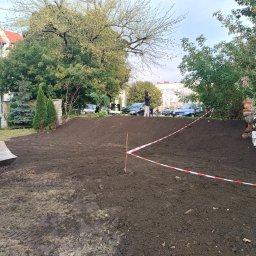 Krzysztof Krankowski - Niwelacja terenu: wyrównana ziemia z wykopem, zabezpieczona taśmą ostrzegawczą. W tle budynek i zieleń, z boku widoczny fragment koparki.