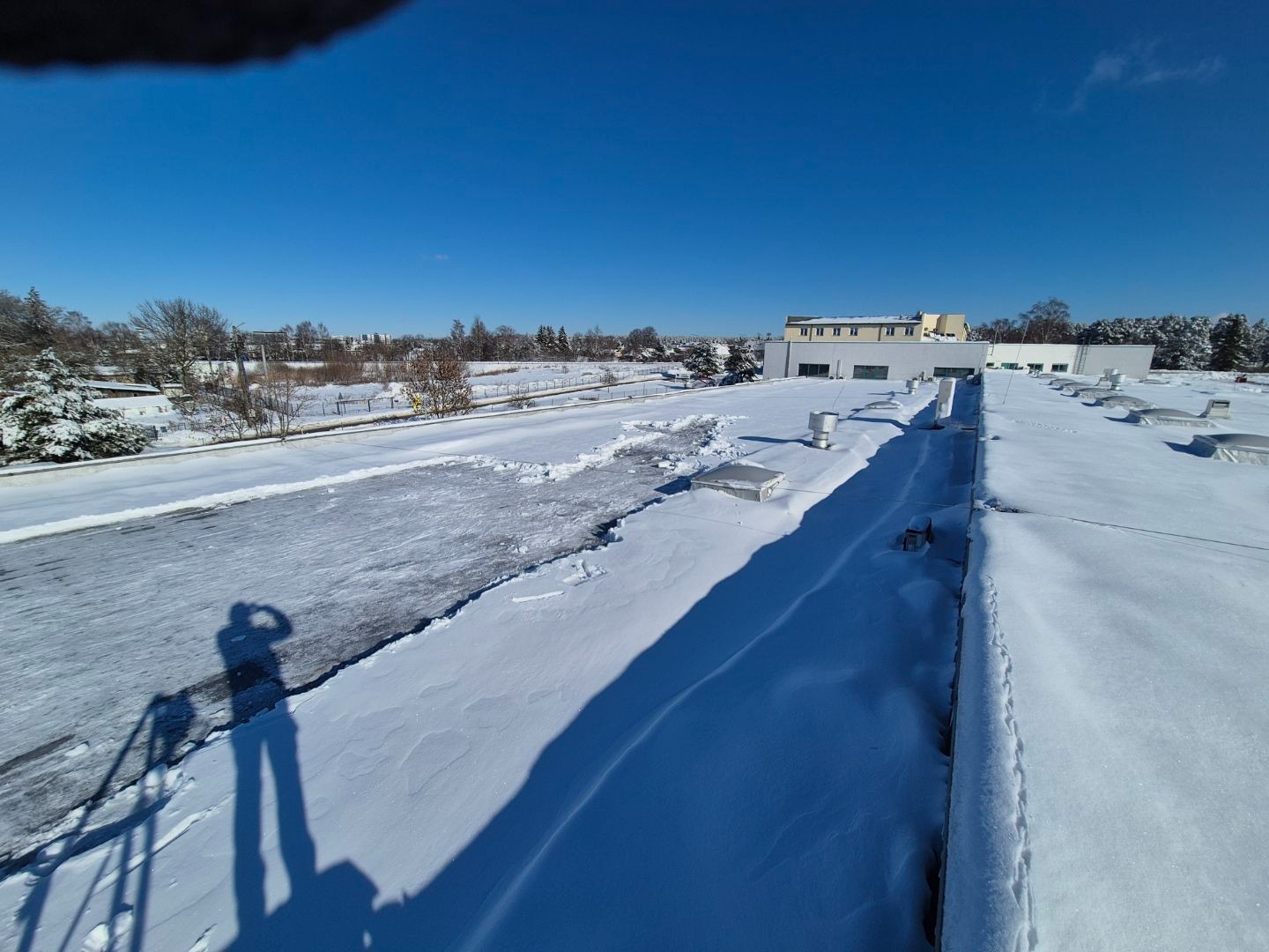 Częściowo odśnieżony dach budynku w słoneczny dzień. Widoczny cień osoby robiącej zdjęcie. Śnieg zalega na pozostałej części dachu i w tle.