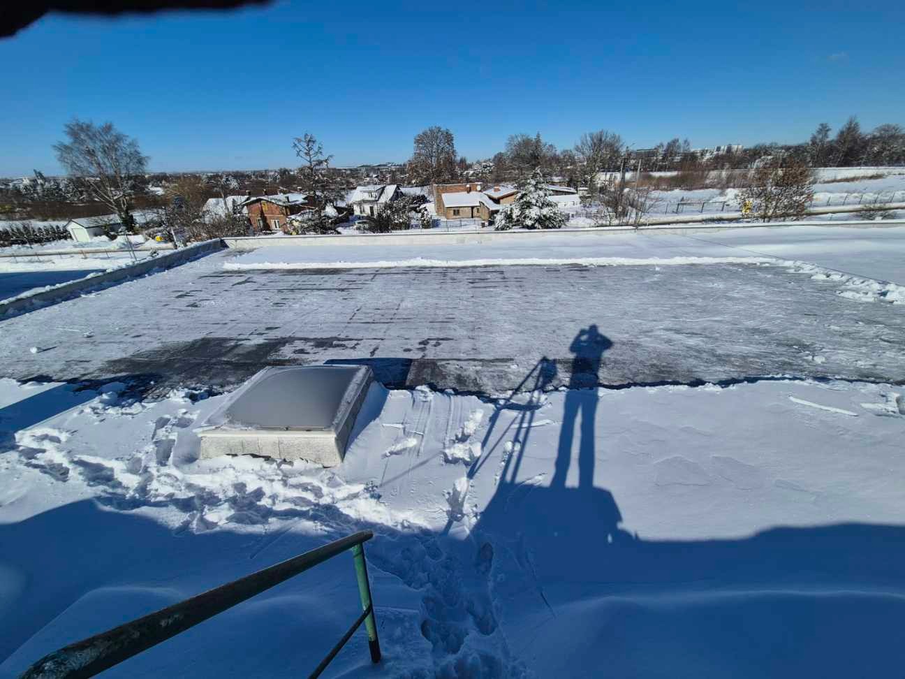 Śnieg na dachu budynku z świetlikiem, cień osoby na śniegu, widok na zasypane śniegiem domy i drzewa w oddali, błękitne niebo.