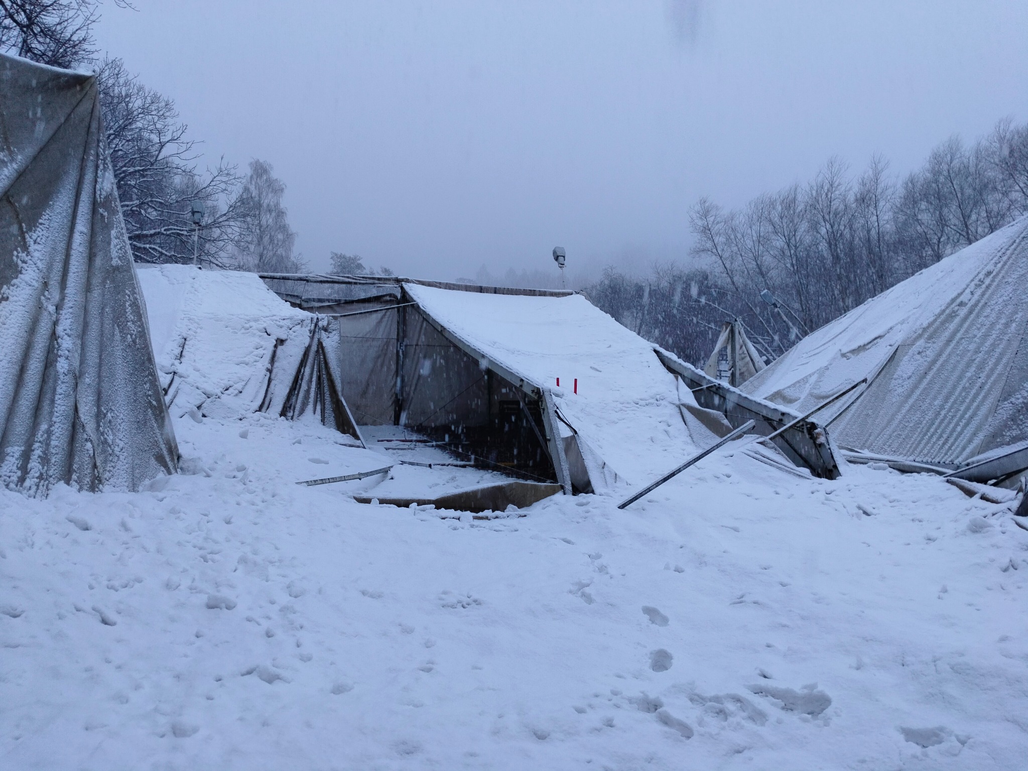 Zasypana śniegiem konstrukcja, prawdopodobnie namiotowa, z widocznymi elementami metalowymi i materiałowymi. W tle drzewa i szara, zimowa aura.