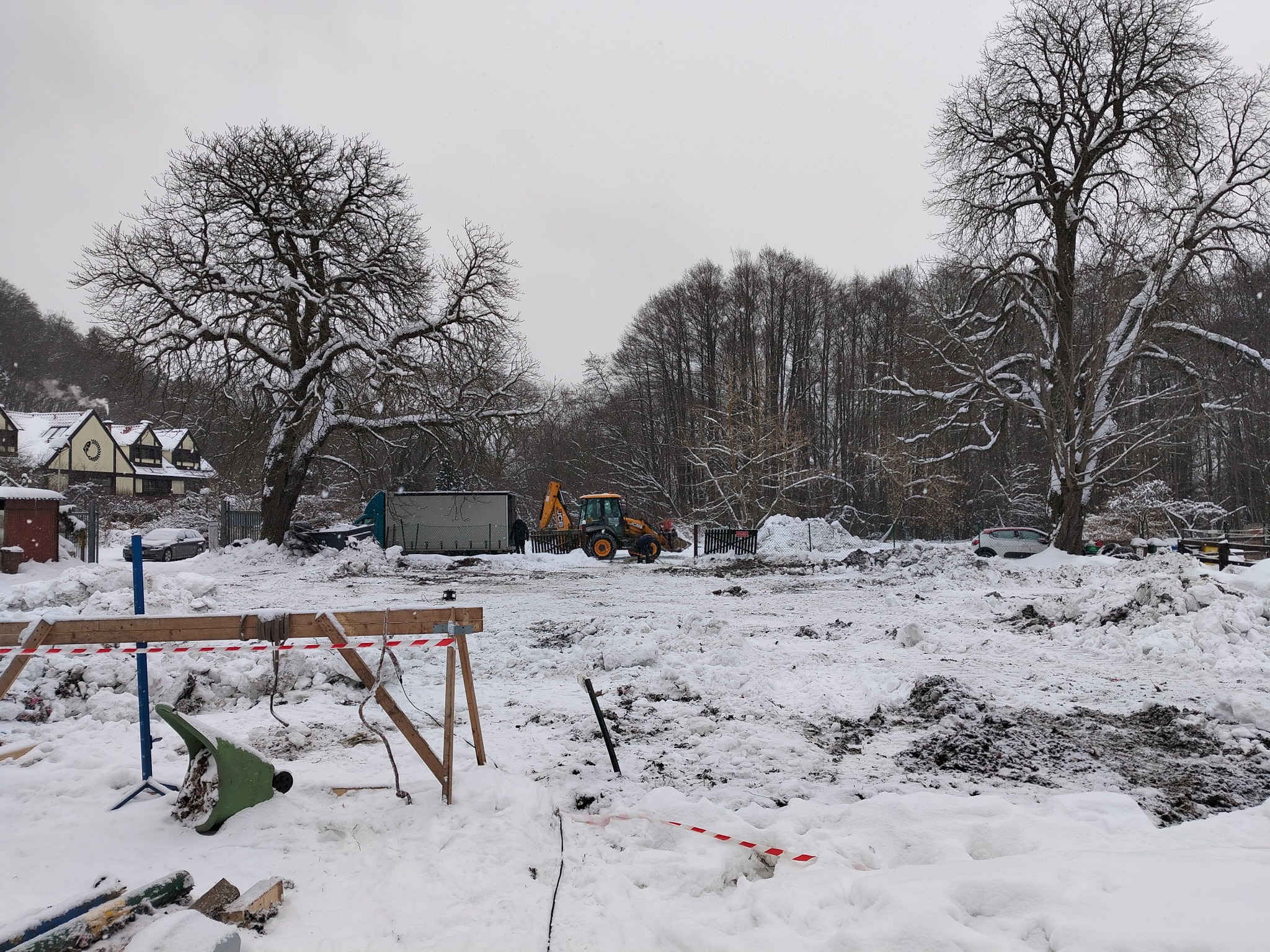 Zimowy krajobraz z wykopem, traktorem i stertami śniegu. Widoczne drzewa bez liści i budynek w tle. Prace ziemne w trakcie odśnieżania.