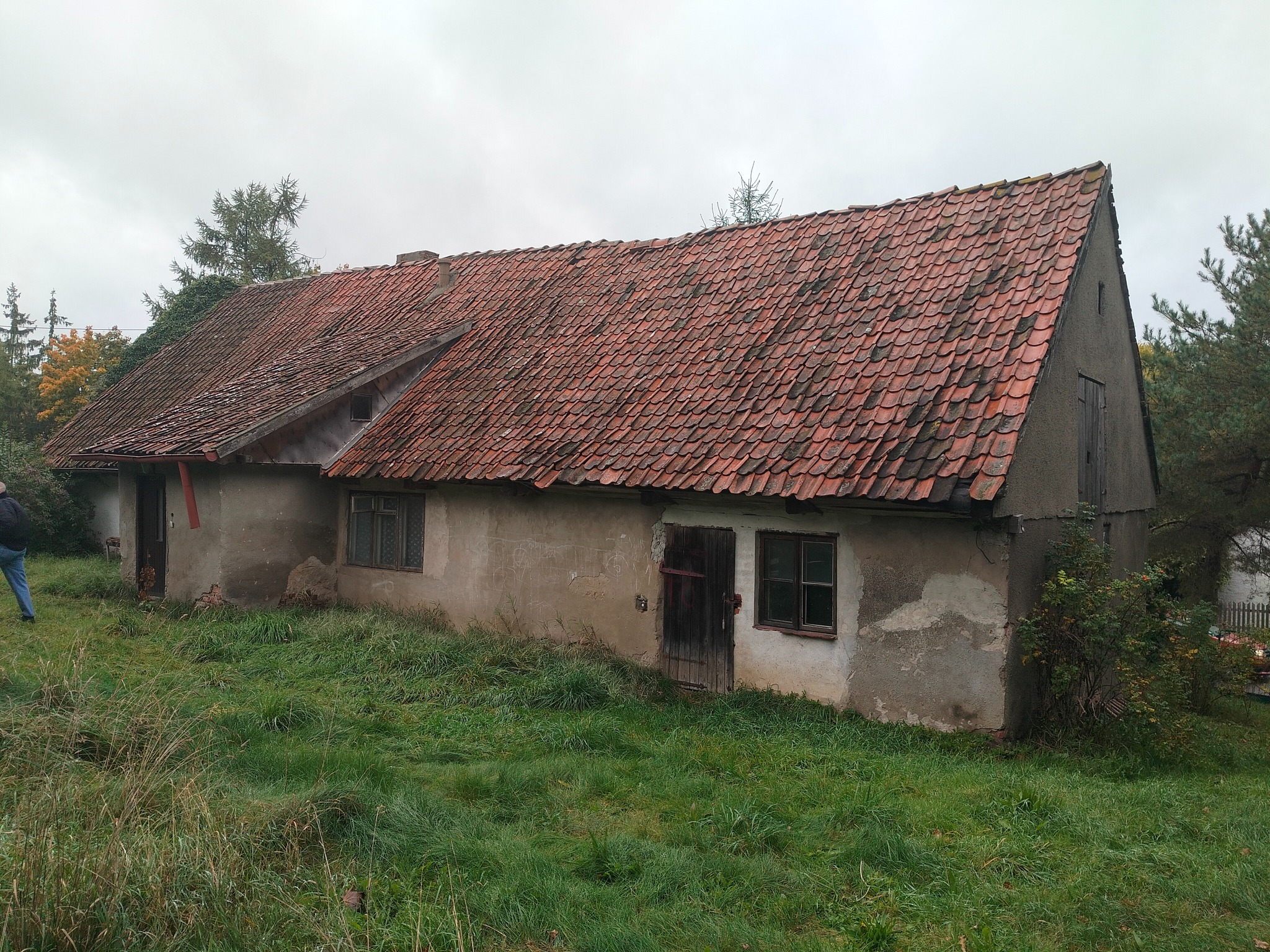 Stary, zaniedbany budynek z czerwoną dachówką, porośnięty trawą. Widoczne okna i drzwi, ściany z ubytkami tynku. Częściowo widoczna osoba po lewej stronie kadru.
