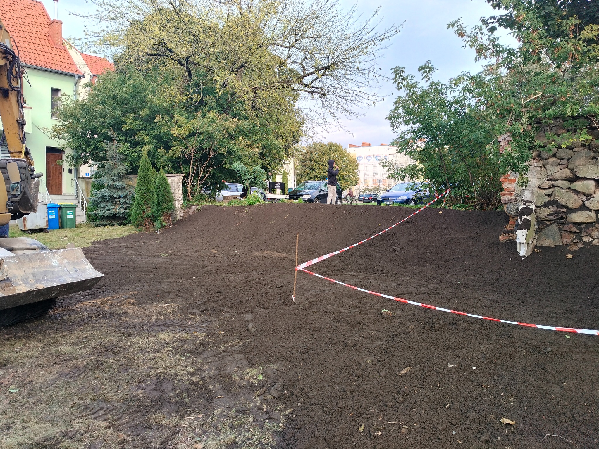 Niwelacja terenu: wyrównana ziemia z wykopem, zabezpieczona taśmą ostrzegawczą. W tle budynek i zieleń, z boku widoczny fragment koparki.