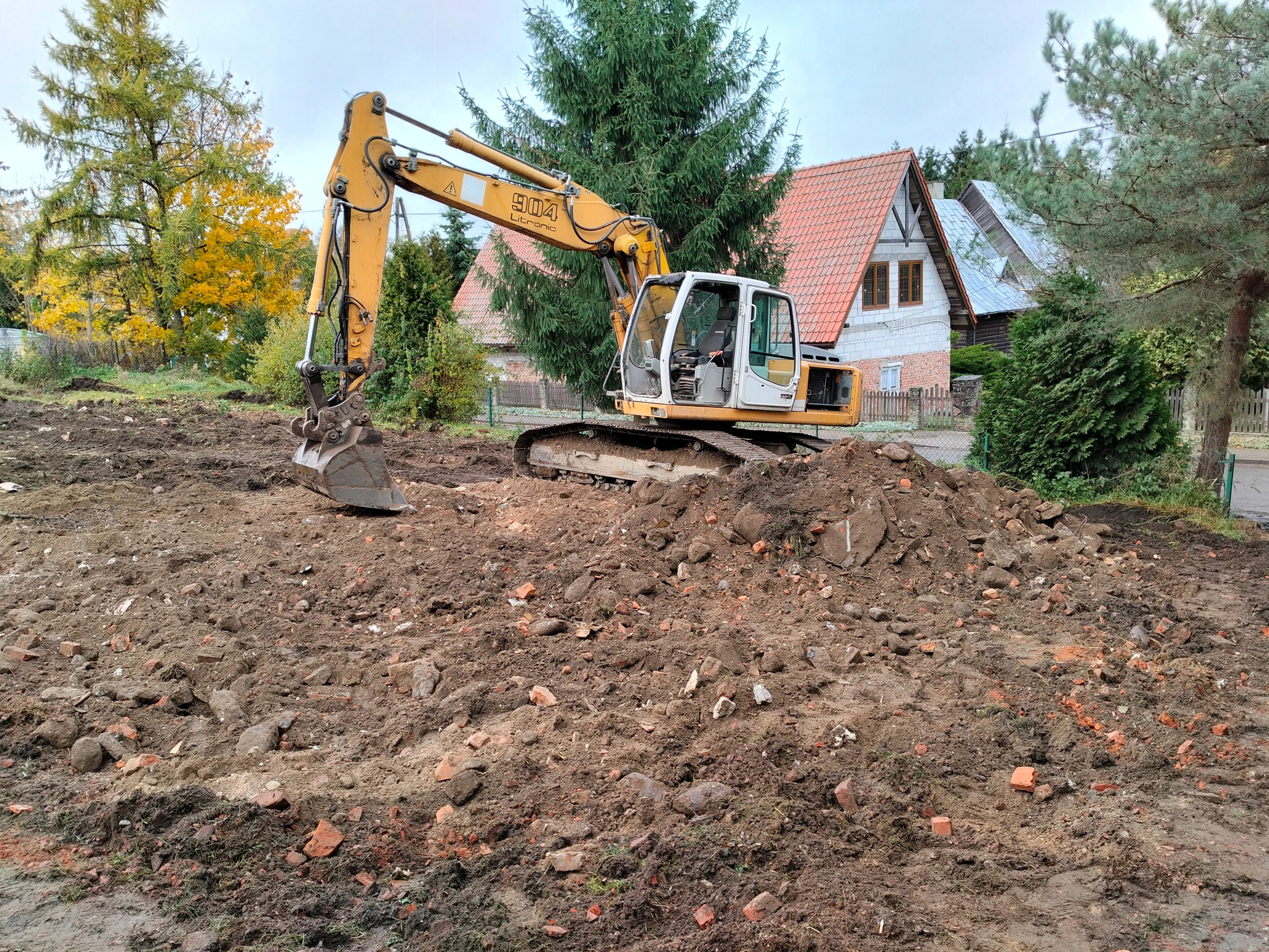 Żółta koparka Litronic 904 na placu budowy z gruzem i ziemią, w tle domy z czerwoną dachówką i drzewa w jesiennych barwach. Prace ziemne w toku.