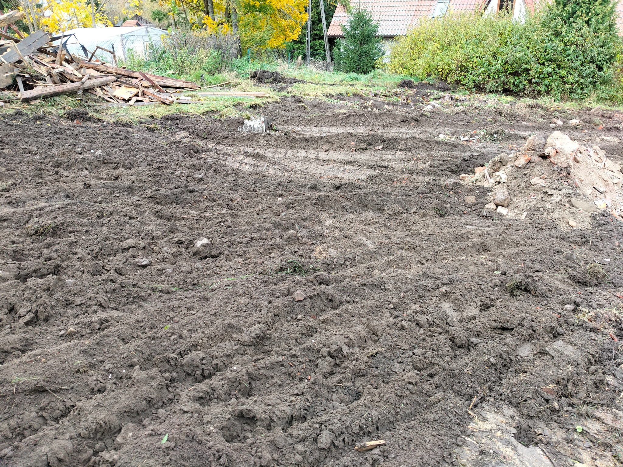 Przygotowany teren po rozbiórce z widocznymi śladami opon, sterta gruzu i resztki drewna w tle. Niwelacja terenu po zakończonych pracach rozbiórkowych.