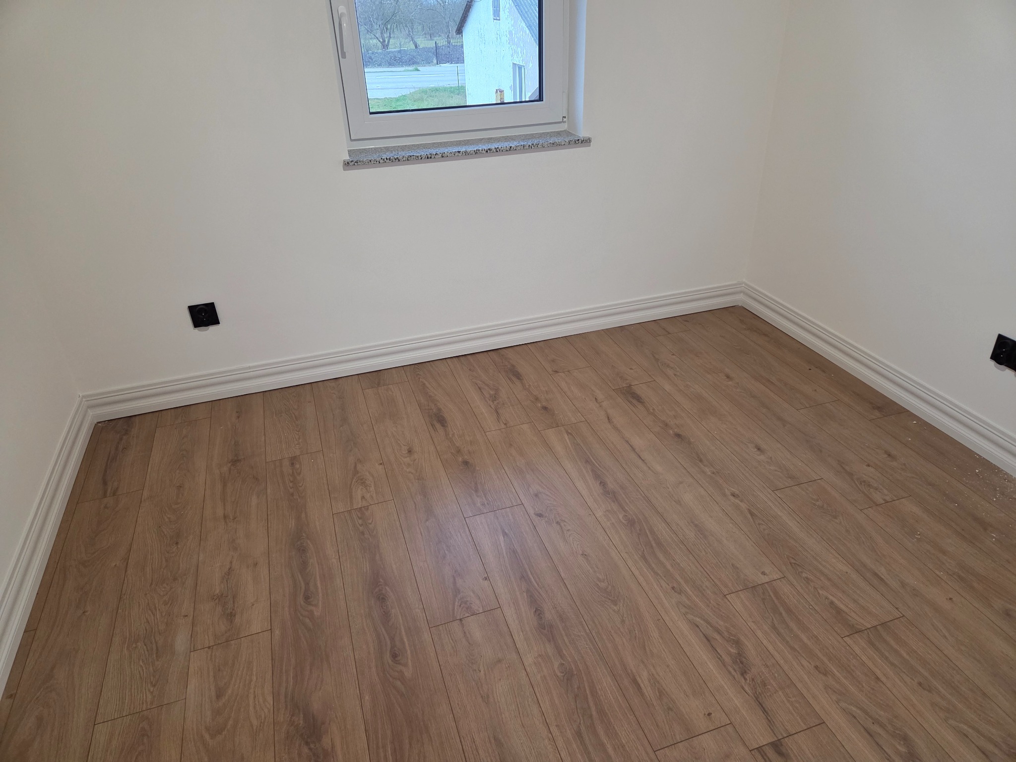 Ułożone panele podłogowe w jasnym pokoju z białymi ścianami i listwami przypodłogowymi. Widoczne gniazdka elektryczne i okno z parapetem.