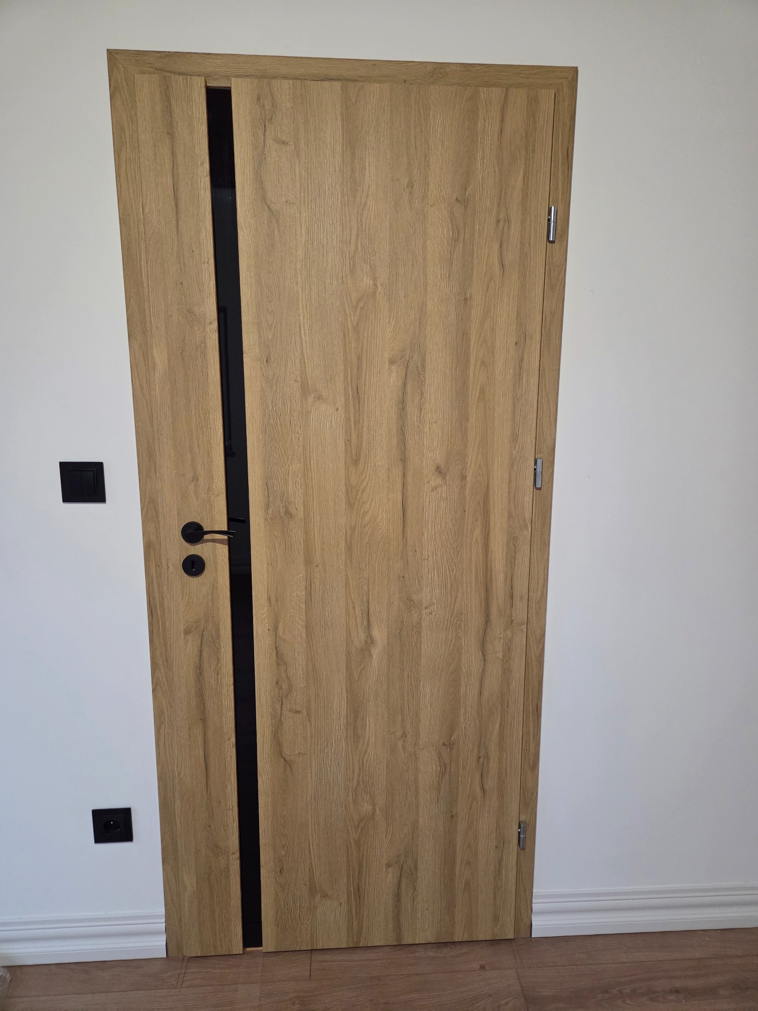 Otwarta, fornirowana drzwi wewnętrzne z czarną ościeżnicą i klamką, widoczne na tle białej ściany z czarnymi włącznikami, na drewnianej podłodze.