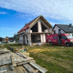 Usługi dekarskie Nowakowski - Budowa domu jednorodzinnego w trakcie krycia dachu czerwoną dachówką. Na dachu pracownik, obok rusztowanie i samochód dostawczy z logo firmy dekarskiej.